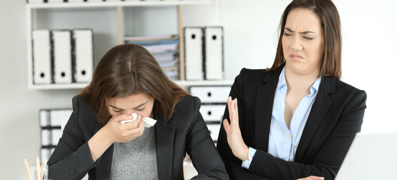 Professional woman sneezing into a tissue and coworker looking with disgust