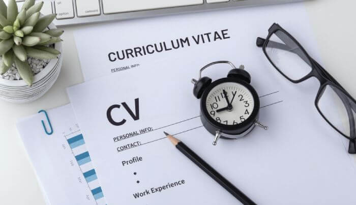 Top view of a desk with a CV, a keyboard, a pencil, a small clock, and reading glasses.